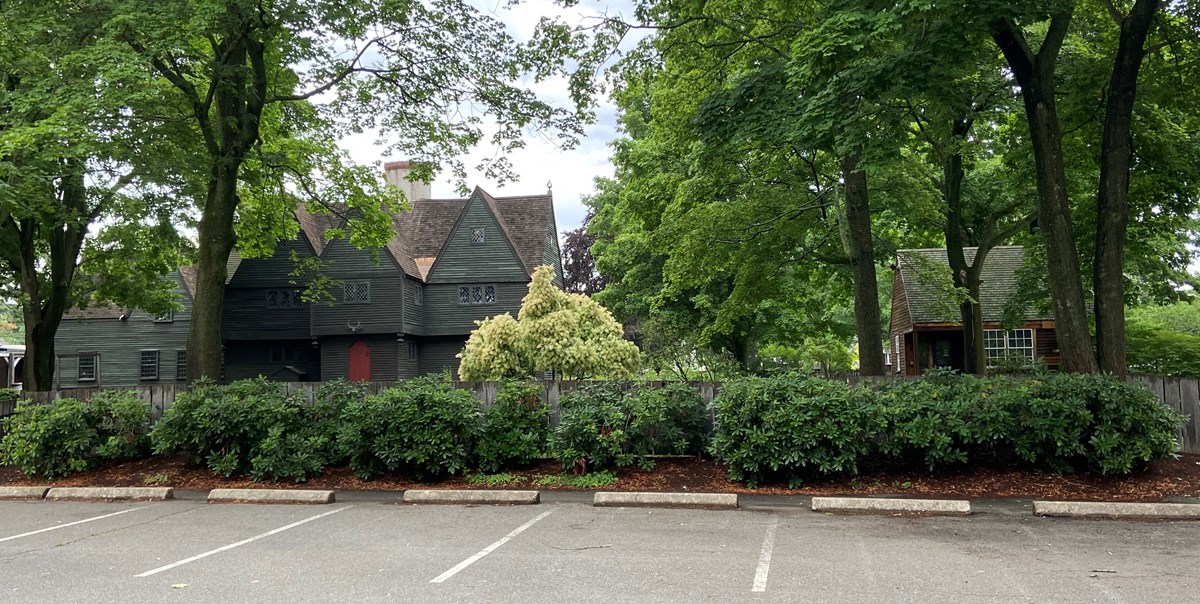 Paved parking area bordered by green trees and shrubs in front of a multi-story historic home.