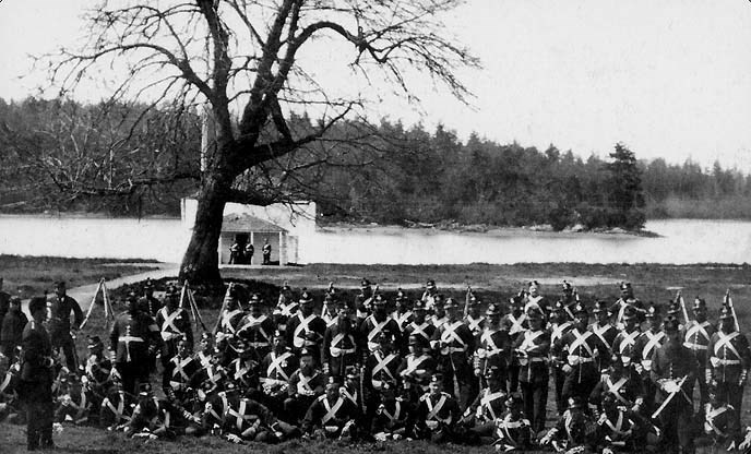 A black and white photo of many English soldiers by a tree. There is a bay in the background and a white small building