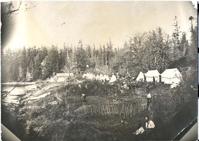 tintype photograph with a wooden palisade at center with a man working on a garden. In the foreground are two more men with tools. Behind the men are 9 white  tents surrounded by trees.  To the left is a body of water with a canoe resting on the ground.