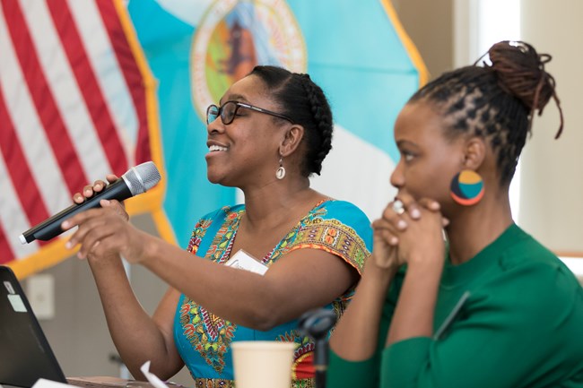 Upper profiles of two women. Woman to the left smiles and speaks into a microphone.