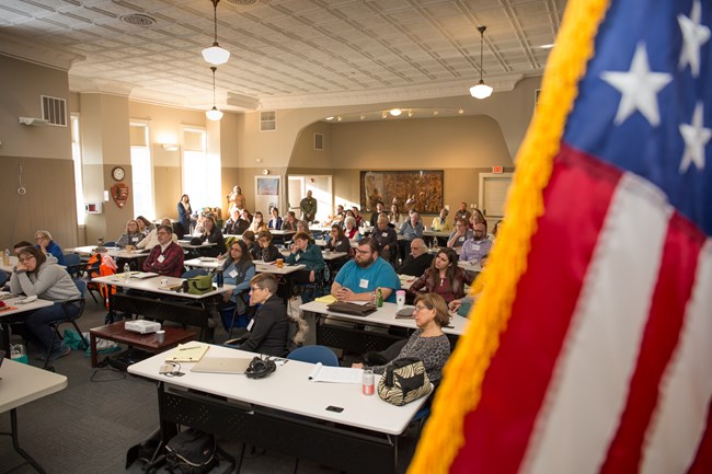 Large room full of people sitting at tables and facing forward. Stars and stripes from the American flag out of focus in foreground.