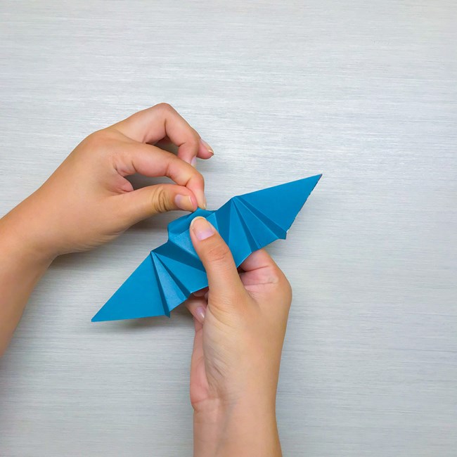 The left index finger and thumb pinching the top corners of the paper to form the bat ears while the right hand holds the bat.