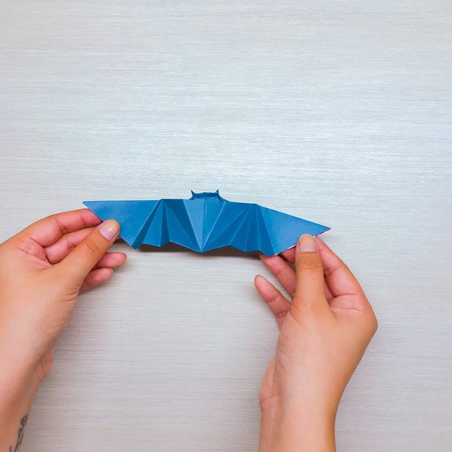 Both hands holding the wings of the completed origami bat flat against the table.