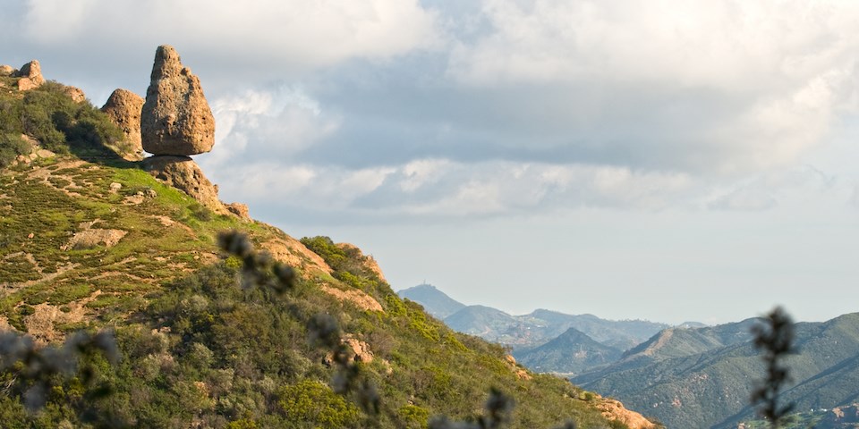 A large rock balances precariously on a cliff while mountain ridges fade into the horizon