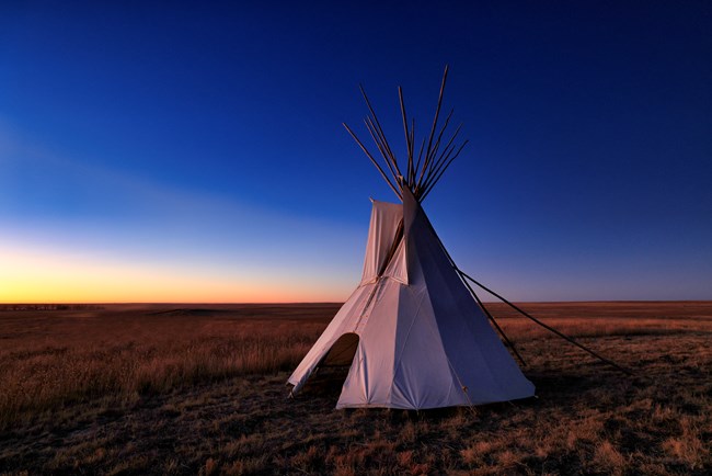 A tipi stands on a prairie landscape in predawn lighting.