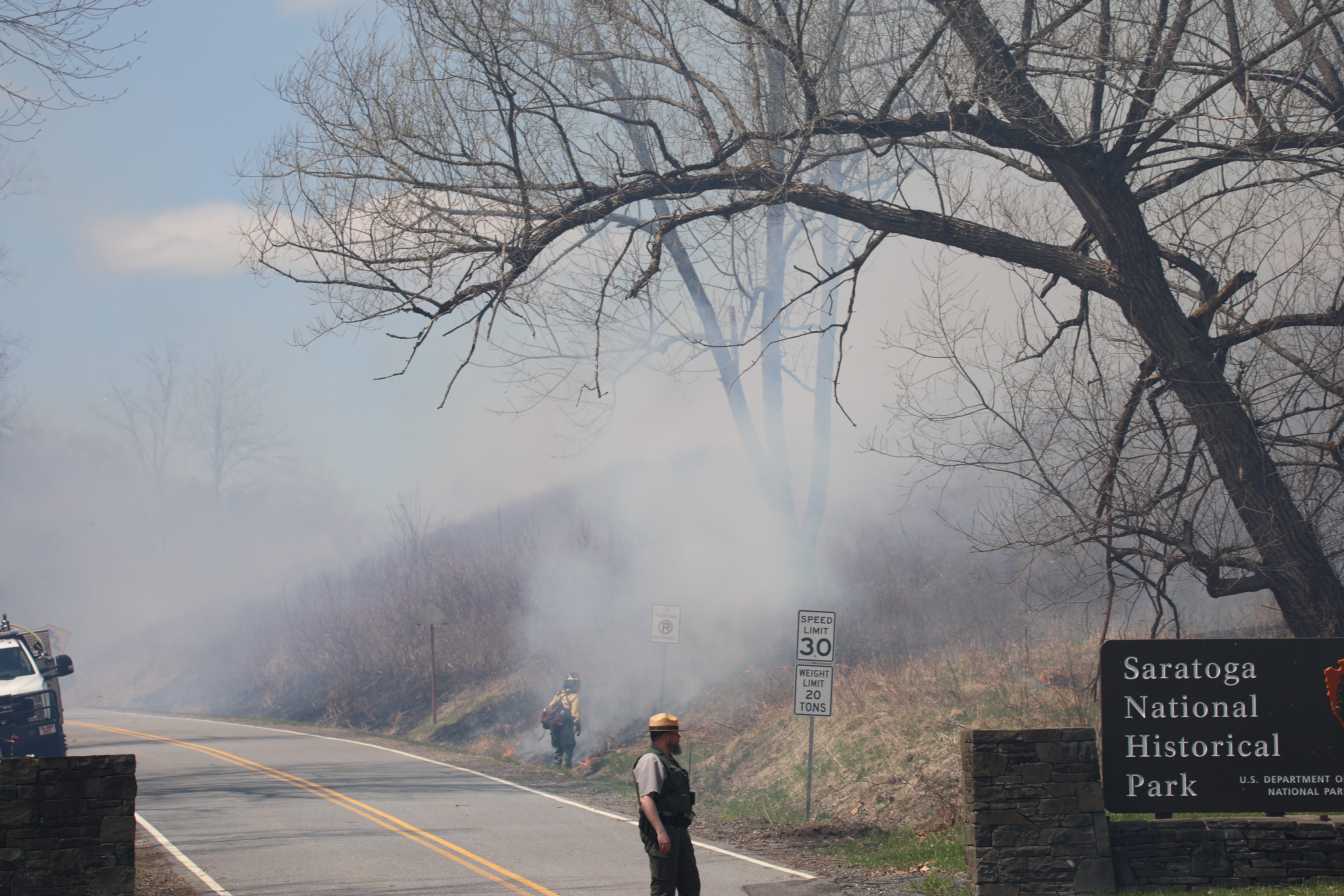 A road runs from the foreground into the distance on the left side of the image. A Park Ranger is conducting trafic with smoke in the background. A firefighter is tending the fire in the midground.