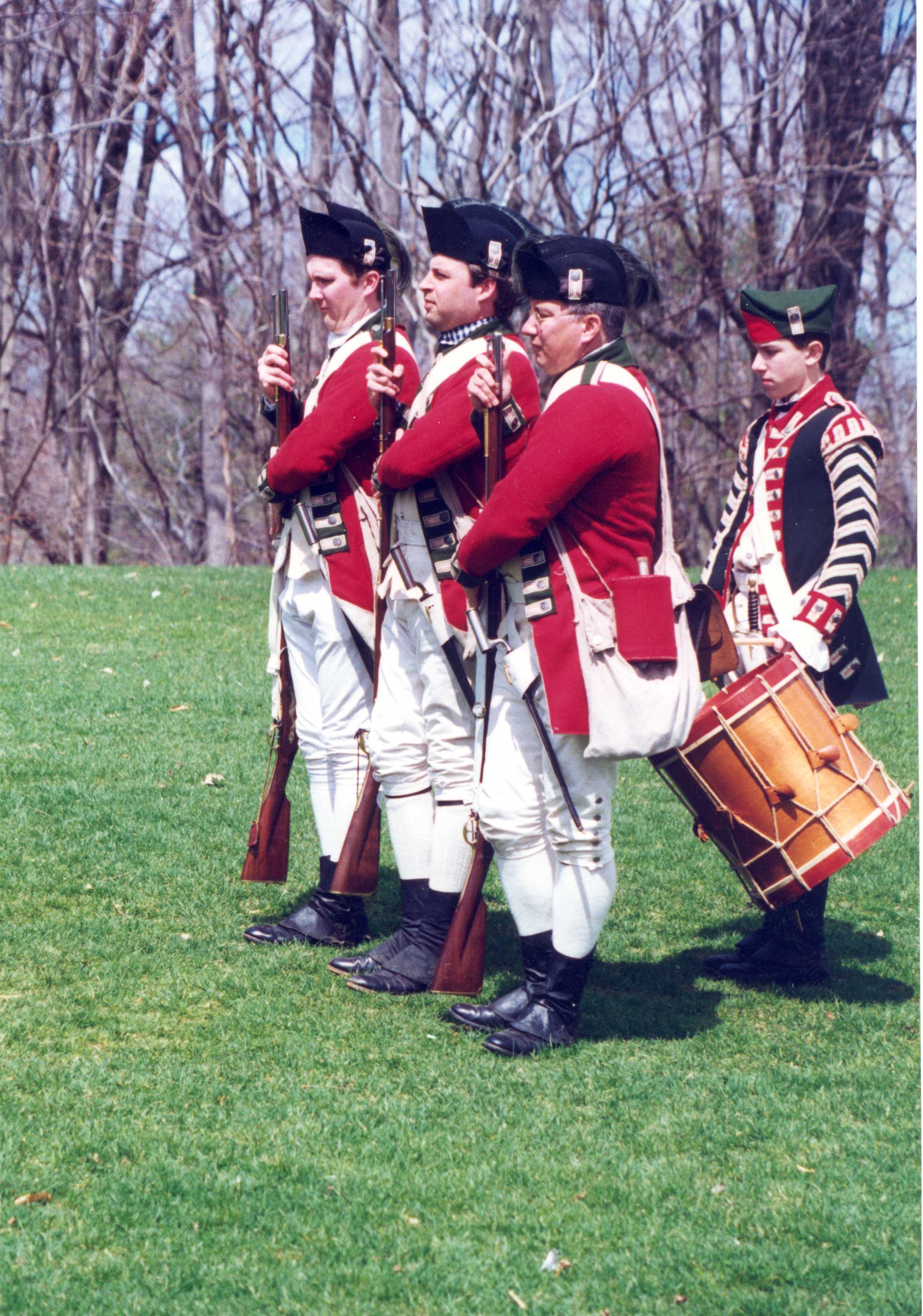3 men in British uniforms march in line as a drummer follows behind