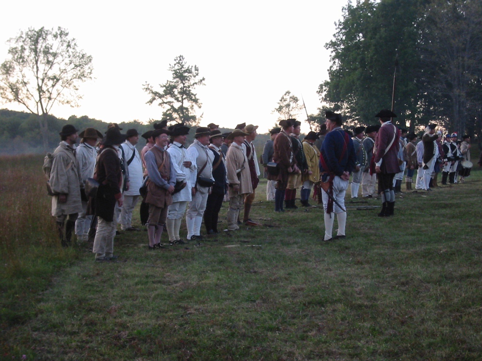 A line of American soldiers walk across a grassy field in a line, shoulder to shoulder, weapons at the ready.