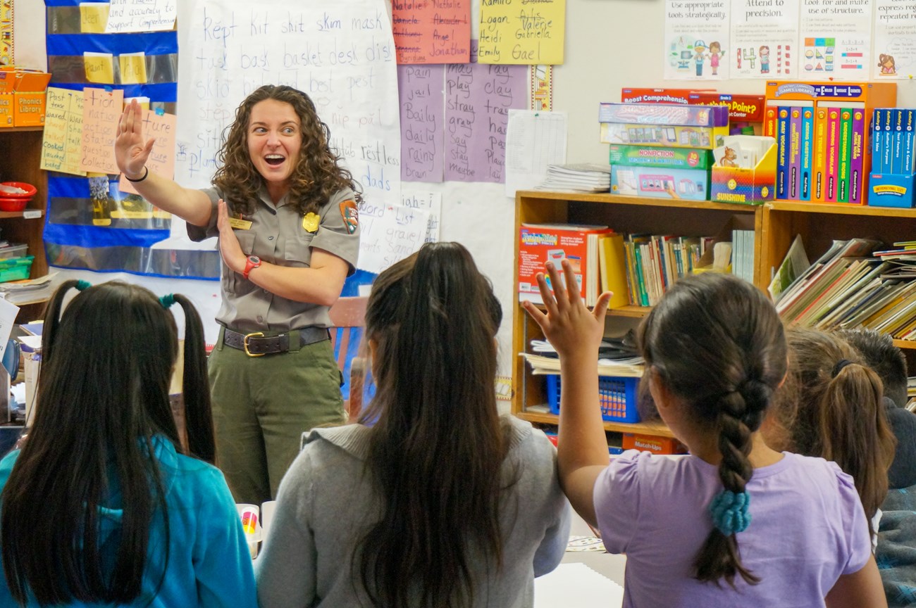 In a classroom, a park ranger lifts her hand in the "stop" position as students watch. One student raises her hand.