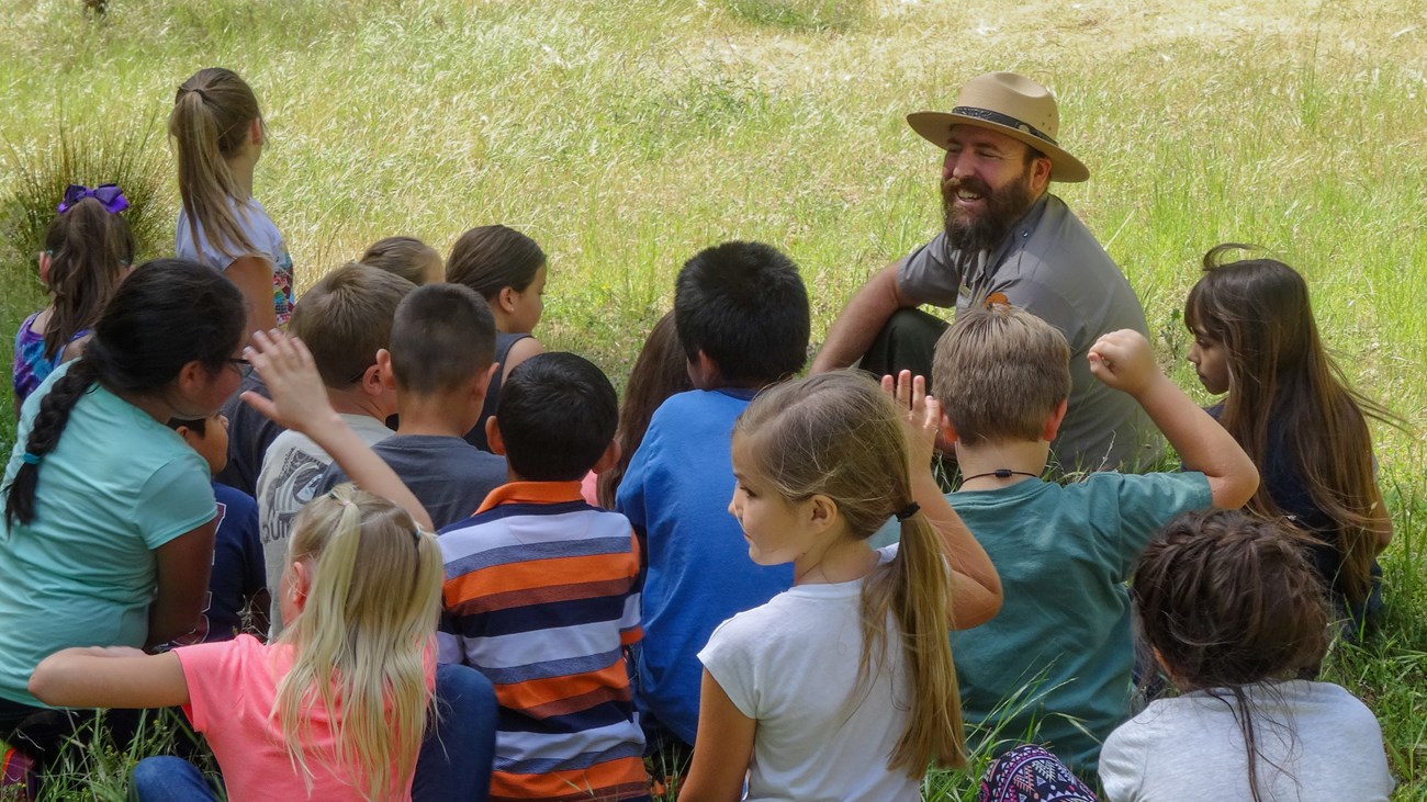 A bearded park ranger sits in a green field with young students crowded around him with their hands raised.