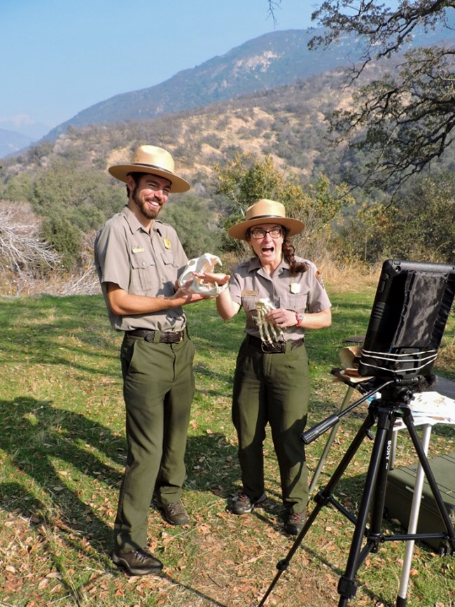 Two park rangers stand outside holding a skull. They are facing a computer tablet on a tripod.