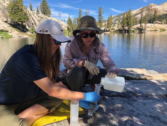 Two women sit at edge of a lake and pour a water sample into a tube to be filtered and collected.