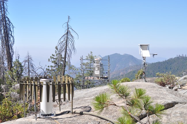 Large granite rock with air quality monitoring equipment and a view of forested mountains and a smoggy valley below.