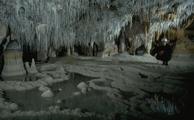 Geologist perches on cave floor near a pool in a room with fragile white formations hanging like icicles from ceiling.