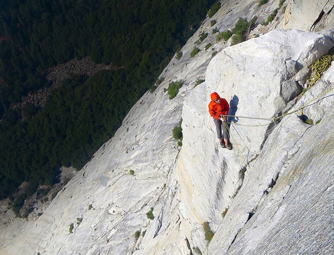 A person roped in and standing on a ledge on the side of a steep granite face