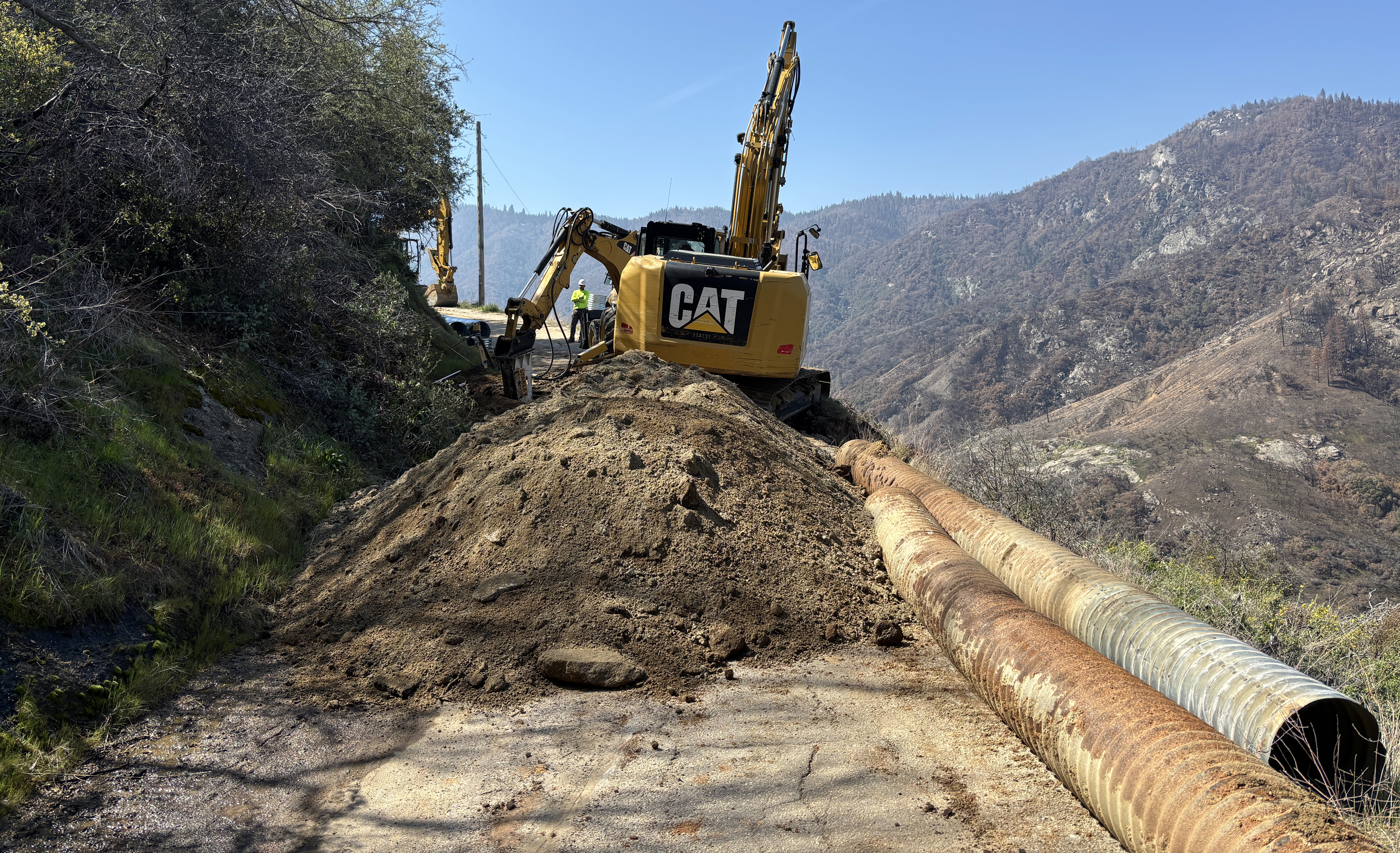 A large pile of dirt next to two long corrugated metal culvert pipes completely block a narrow road in the mountains. A yellow and black piece of construction equipment with the word "Cat" works on the road behind the pile.