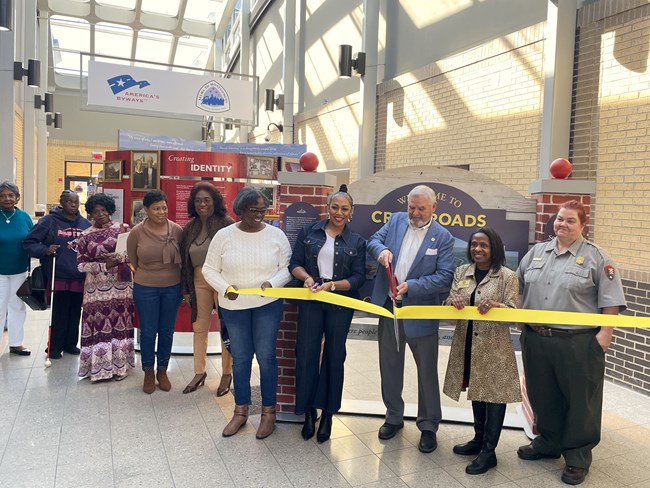 A crowd looks on as several people hold a ribbon which a man uses large decorative scissors to cut.