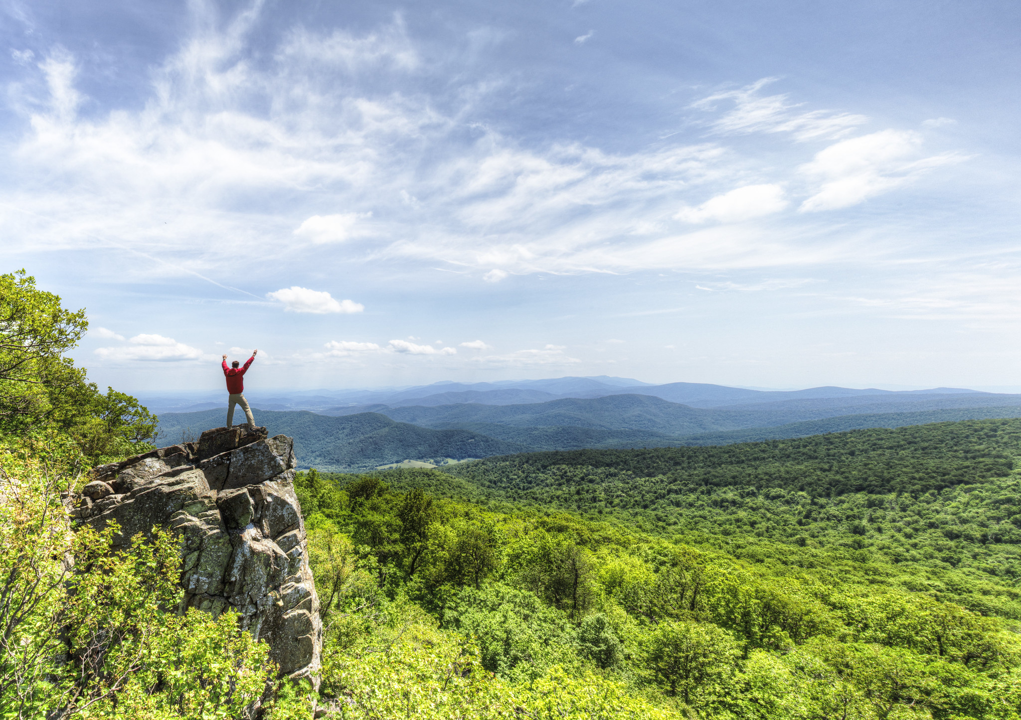A hiker extends his arms in a victorious expression standing on a rock outcrop with a view of the mountain and valley below.
