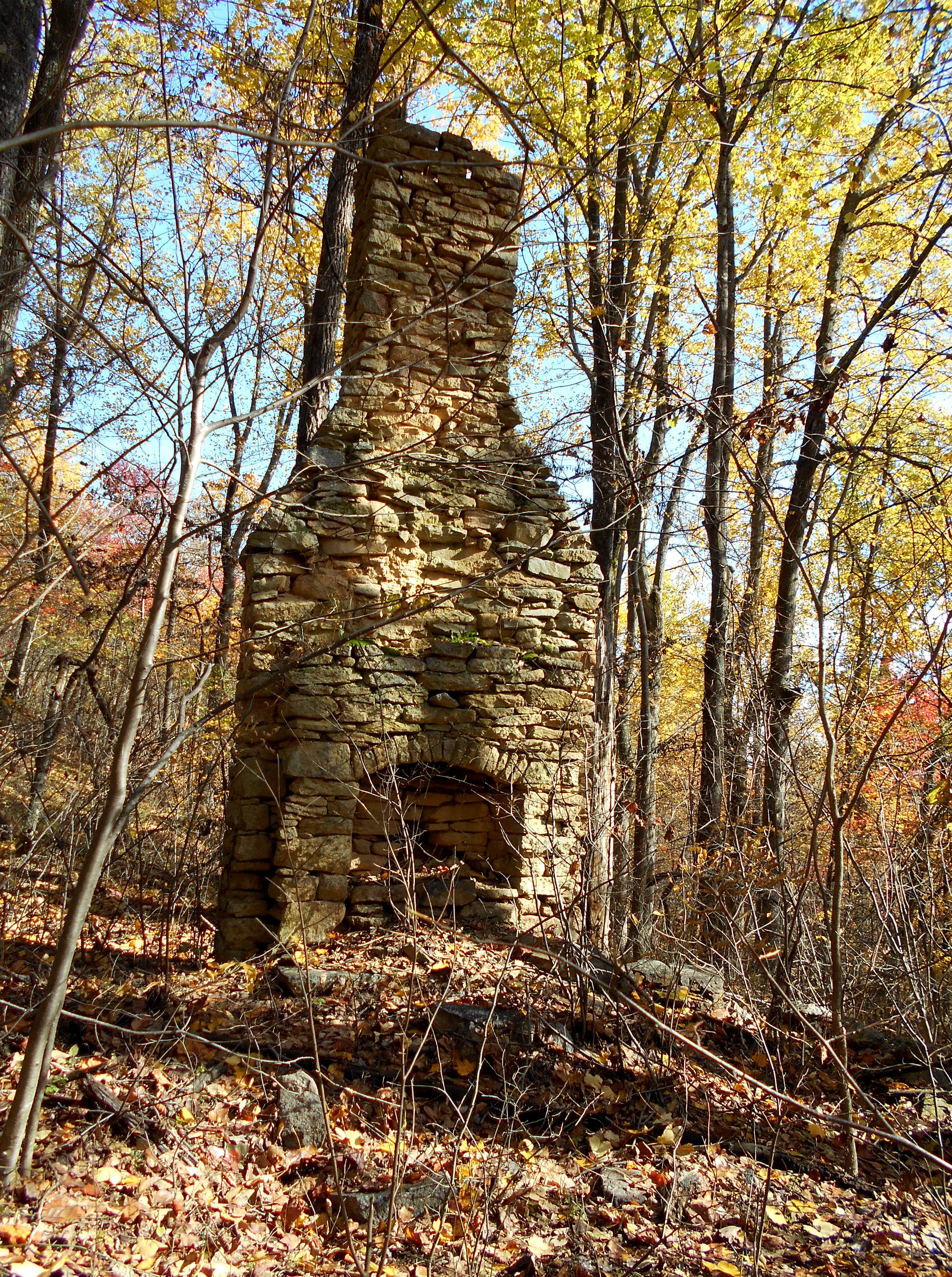 A tan stone chimney stands in the middle of an early autumn forest on a sunny day, leaves of green, yellow, and red decorate the ground