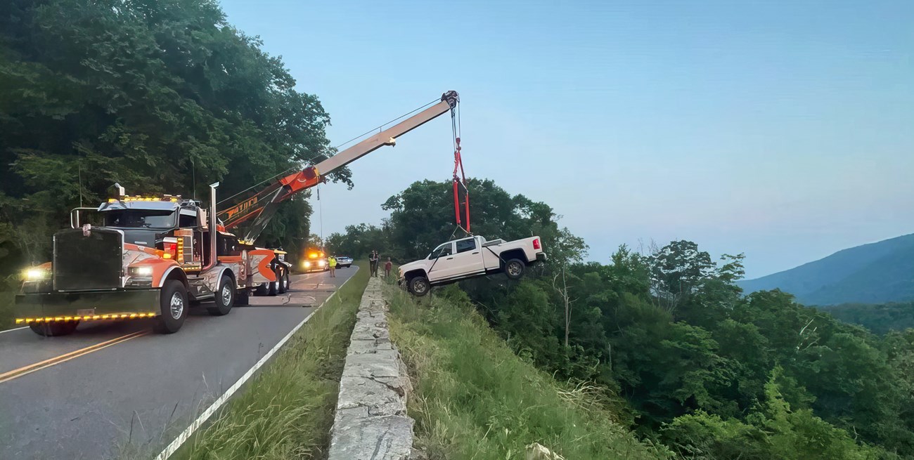 A white pickup truck suspended by a crane. The truck is being recovered after driving off the edge of the mountain road.