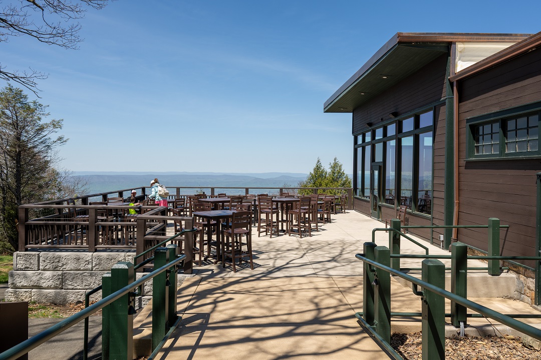 Two people sit at outdoor tables on a concrete patio next to a brown building. In the background, blue rolling mountain peaks under a blue sky. In the foreground is the pathway with green railings, leading to the patio.