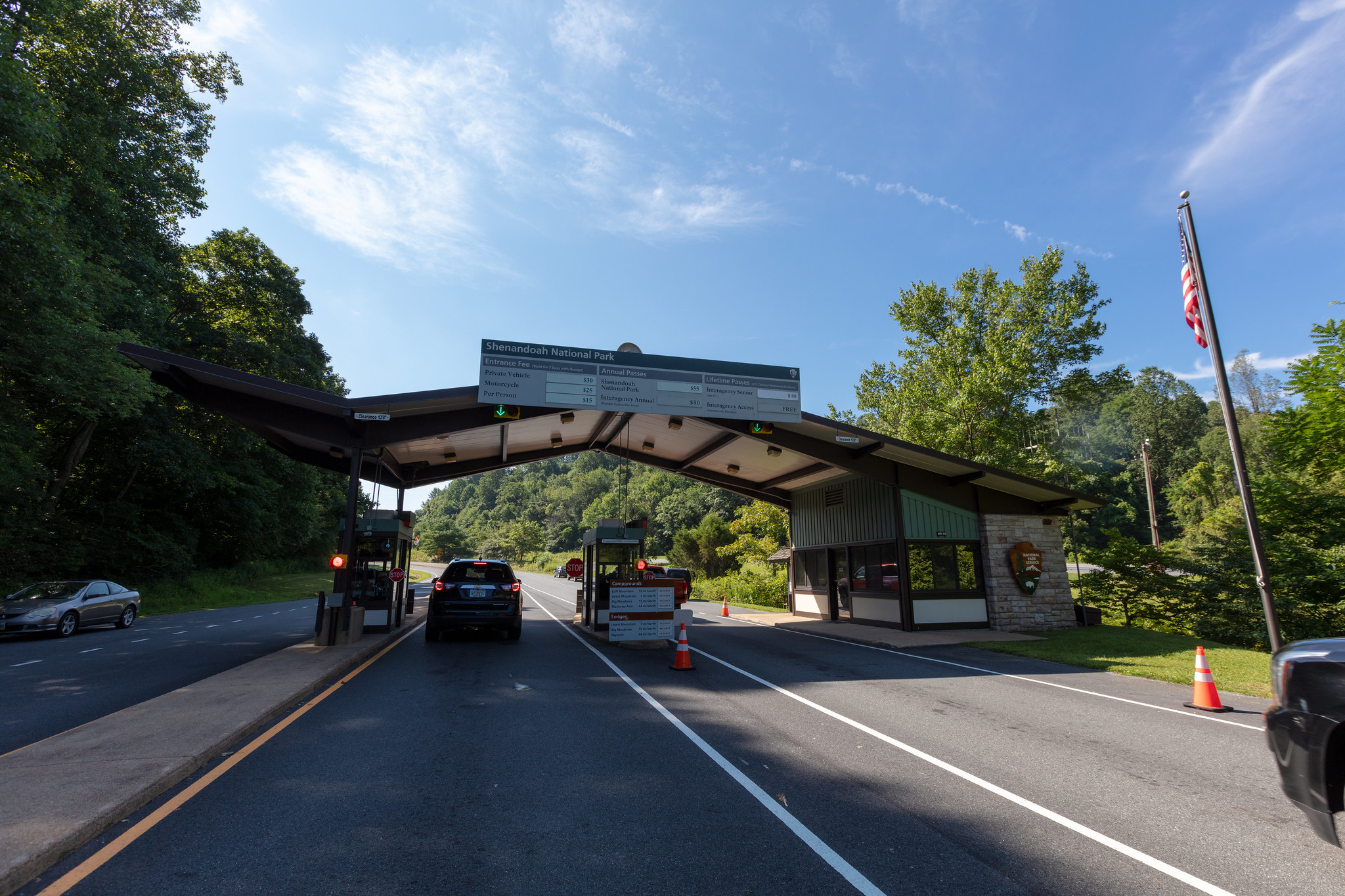A National Park Service entrance station building with two lanes for driving through. A car is stopped under the building at a station. A car exits the park on a road to the left. A U.S. Flag is on a flag pole at right.