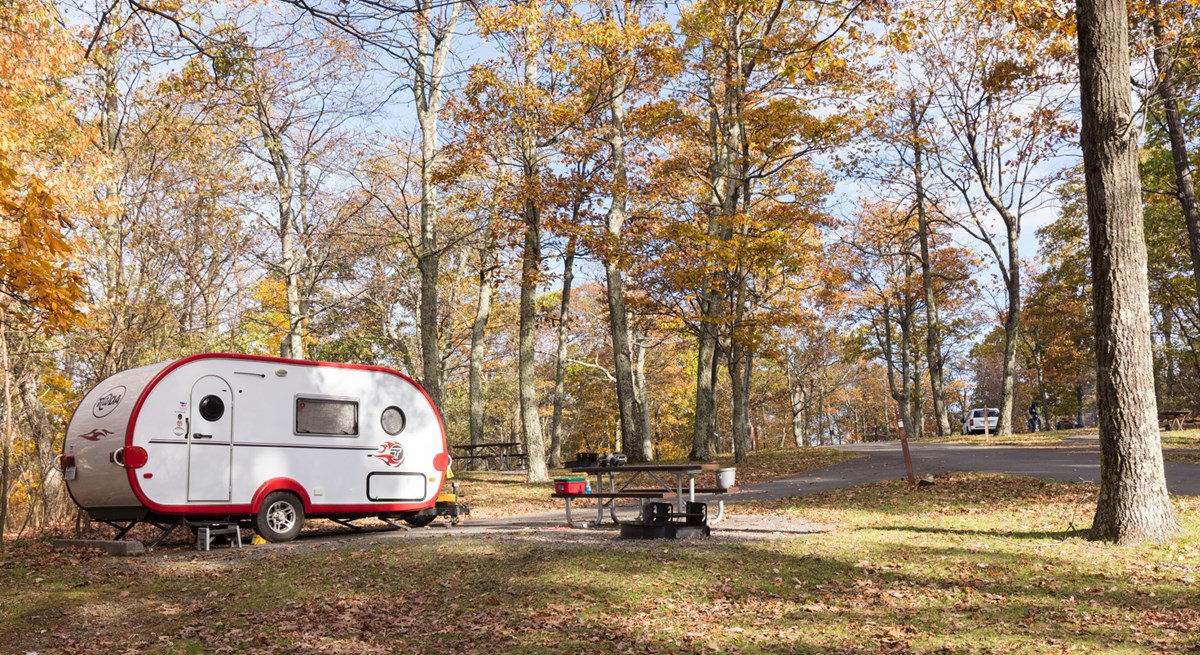 A red and white camper is parked under green trees in a campground.