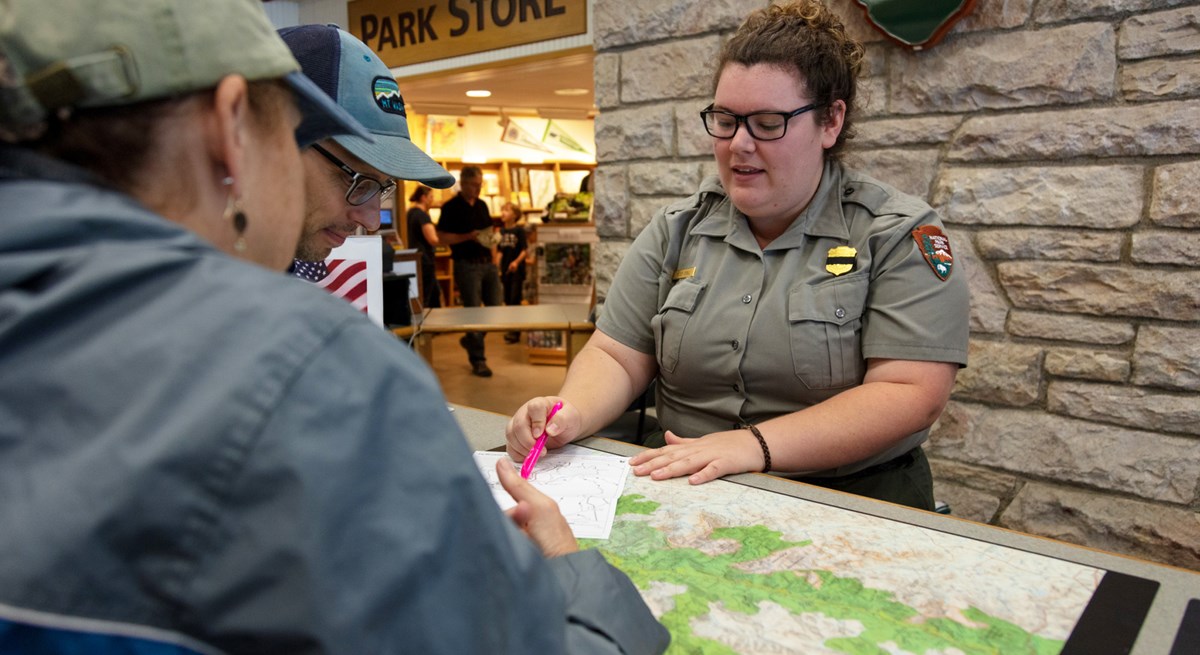 A female park ranger standing behind a desk at a visitor center points to a map for 2 visitors.