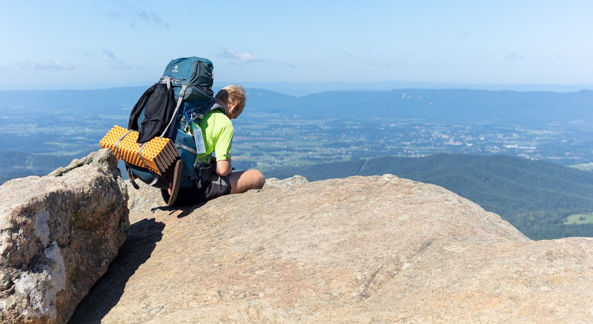 A man with a large backpack sits down on boulders, overlooking a view into the valley below.