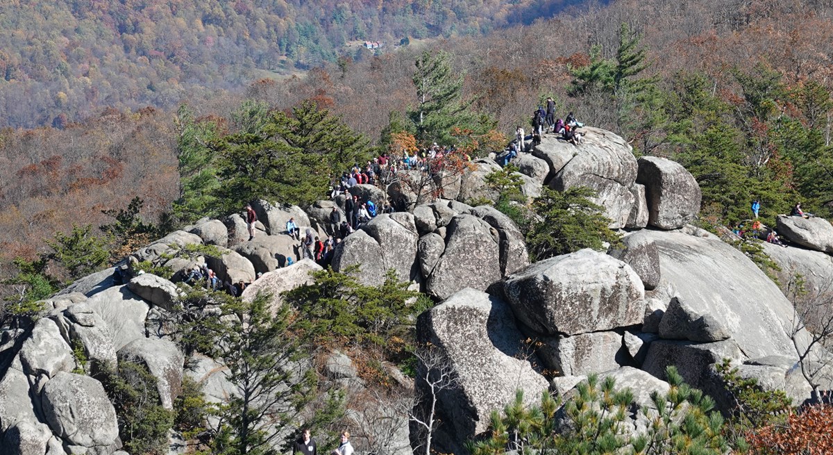 A large crowd of hikers walk along the rocky ridge of a mountain.
