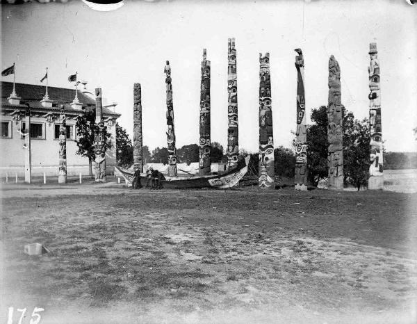 The Waasgo Legend Pole at the 1905 Lewis and Clark Exposition, there are 10 other poles standing beside it, a carved canoe in front of the poles, and the corner of a building in the background. The photo is in black and white.