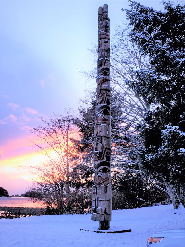 A Totem Pole surrounded by snow. Behind the pole there are trees to the right and a sunset on the ocean on the left.