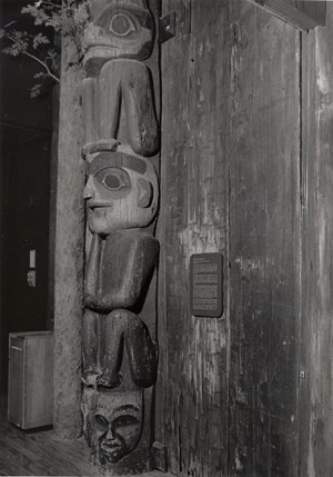 Black and white photograph of the Raven Head Down Pole on display indoors, with a informational plaque hanging next to it.