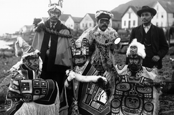 Six Tlingit men, five of whom wear ceremonial garb including headdresses and intricately patterned robes.