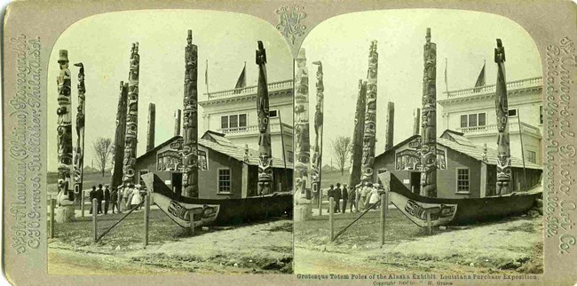 Identical side-by-side black and white photographs of the Alaska exhibit at the 1904 World's Fair, with 6 totems poles, a large canoe and a clan house.