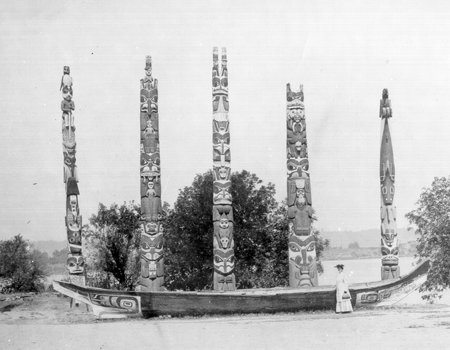 Black and white photograph of five totem poles lined up behind a large carved wood canoe, with a woman in 1900's dress standing on the right end of the canoe.