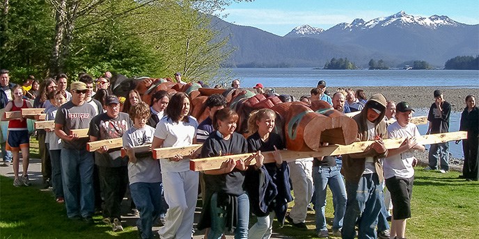 Dozens of youths and adults carrying totem pole onto a grass lawn with ocean and mountains in the background.