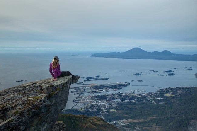 A hiker sits on the edge of the rock at the peak of Verstovia Mountain, overlooking the Sitka Sound.