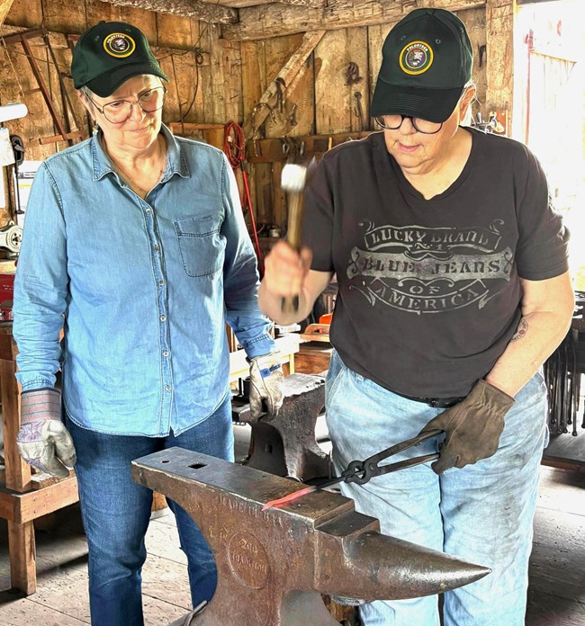 Two volunteer blacksmiths, one overseeing while the other is hammering on an anvil.
