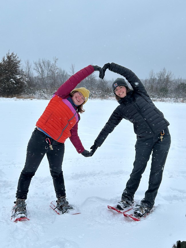 Two people wearing snowshoes hold their arms together to make the shape of a heart.