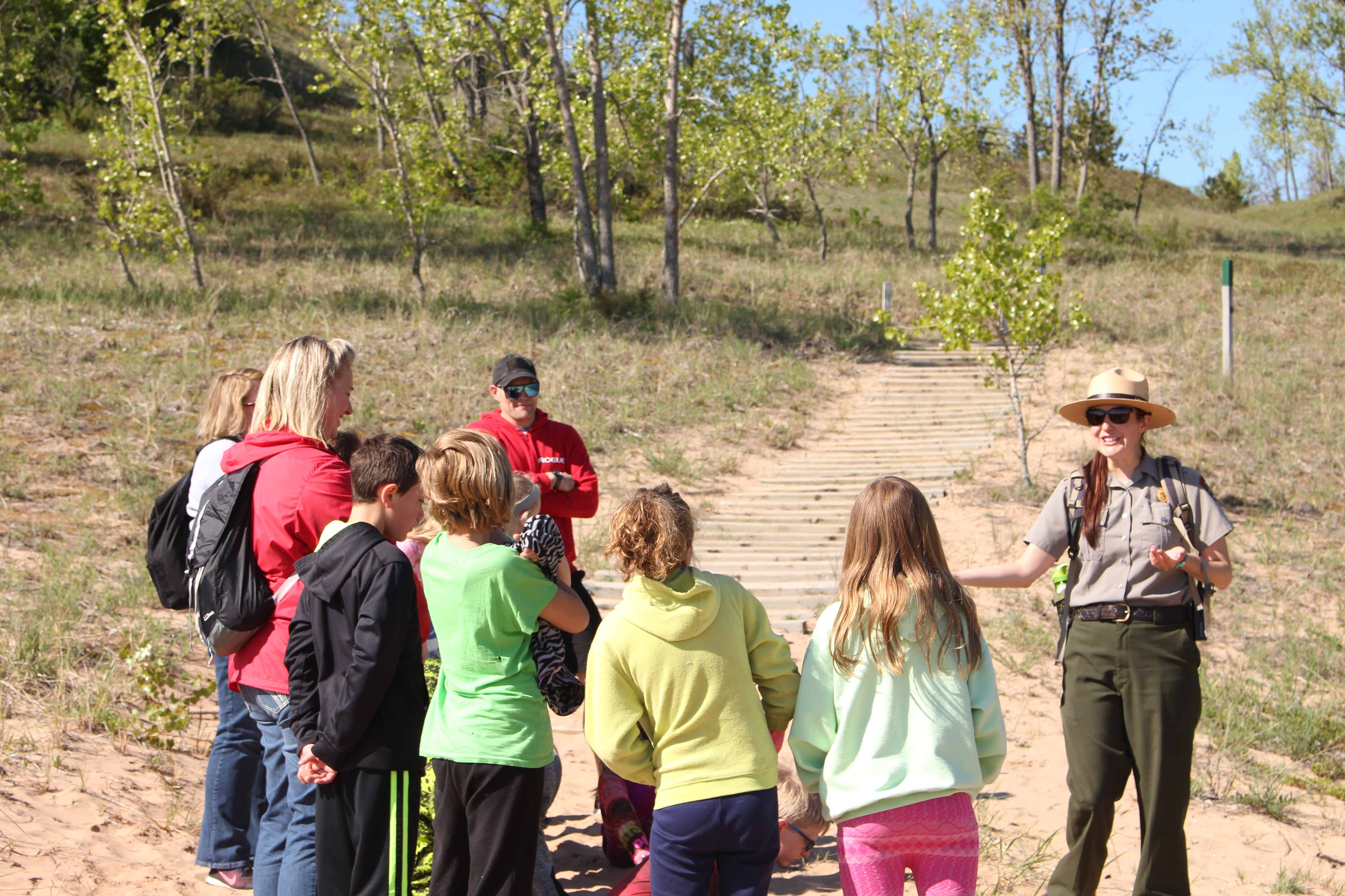 A ranger speaks with a group of students and chaperones standing along a sandy trail lined with grass and trees