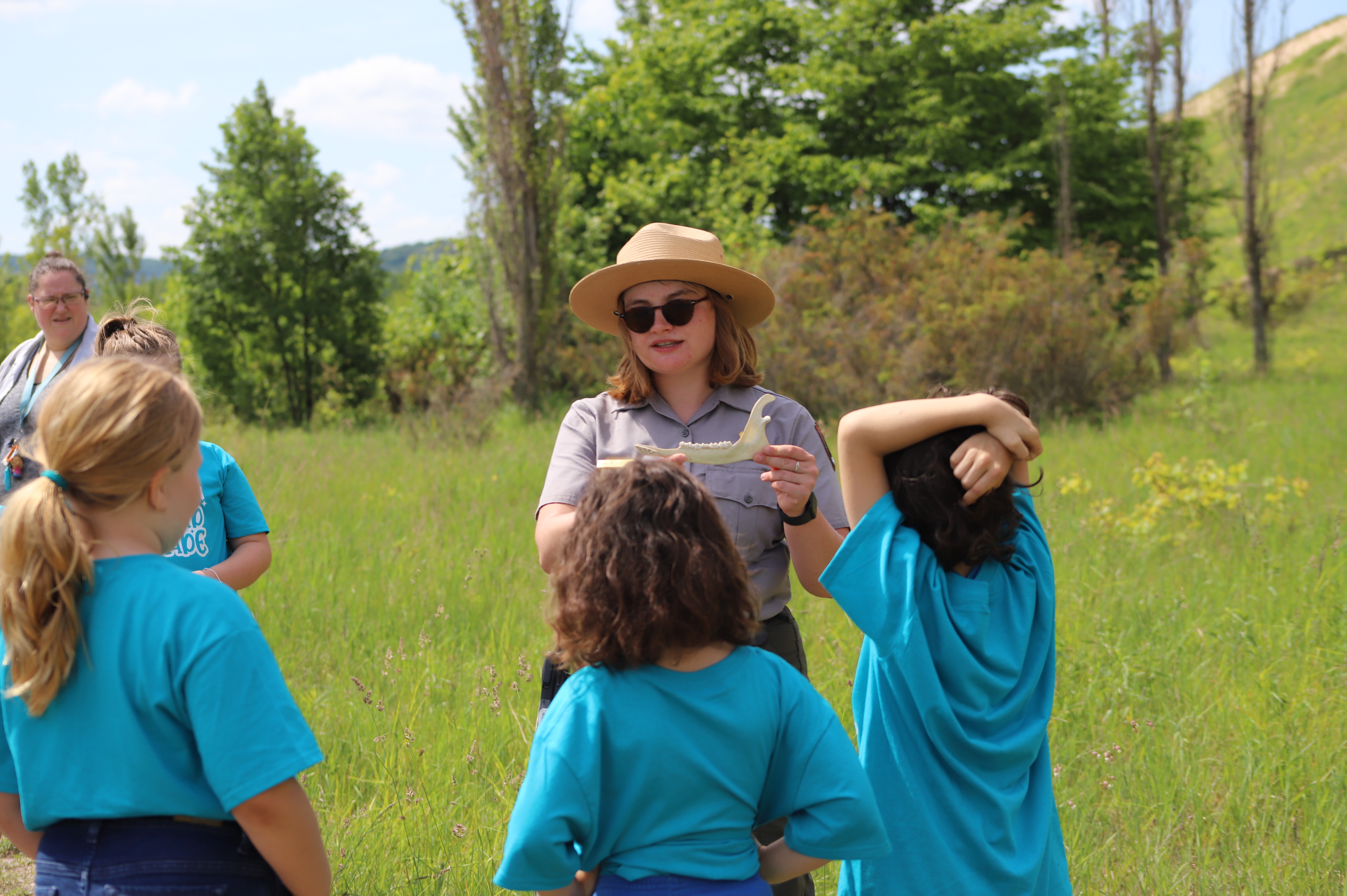 A ranger wearing a flat straw hat and sunglasses displays a deer jawbone to a group of elementary school students in matching blue shirts