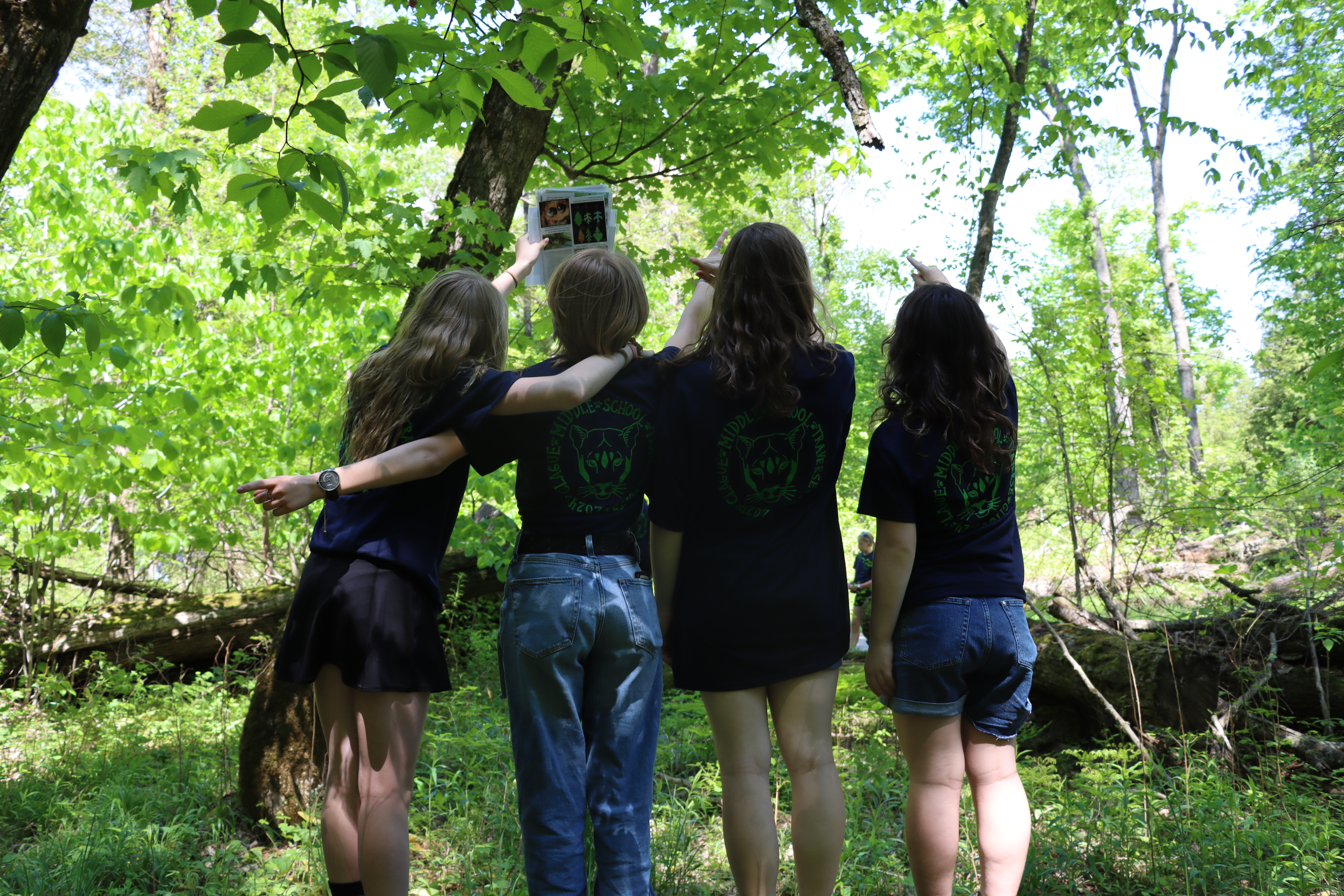 Four middle school students with matching shirts stand together in a green forest, holding an ID sheet up to a tree.