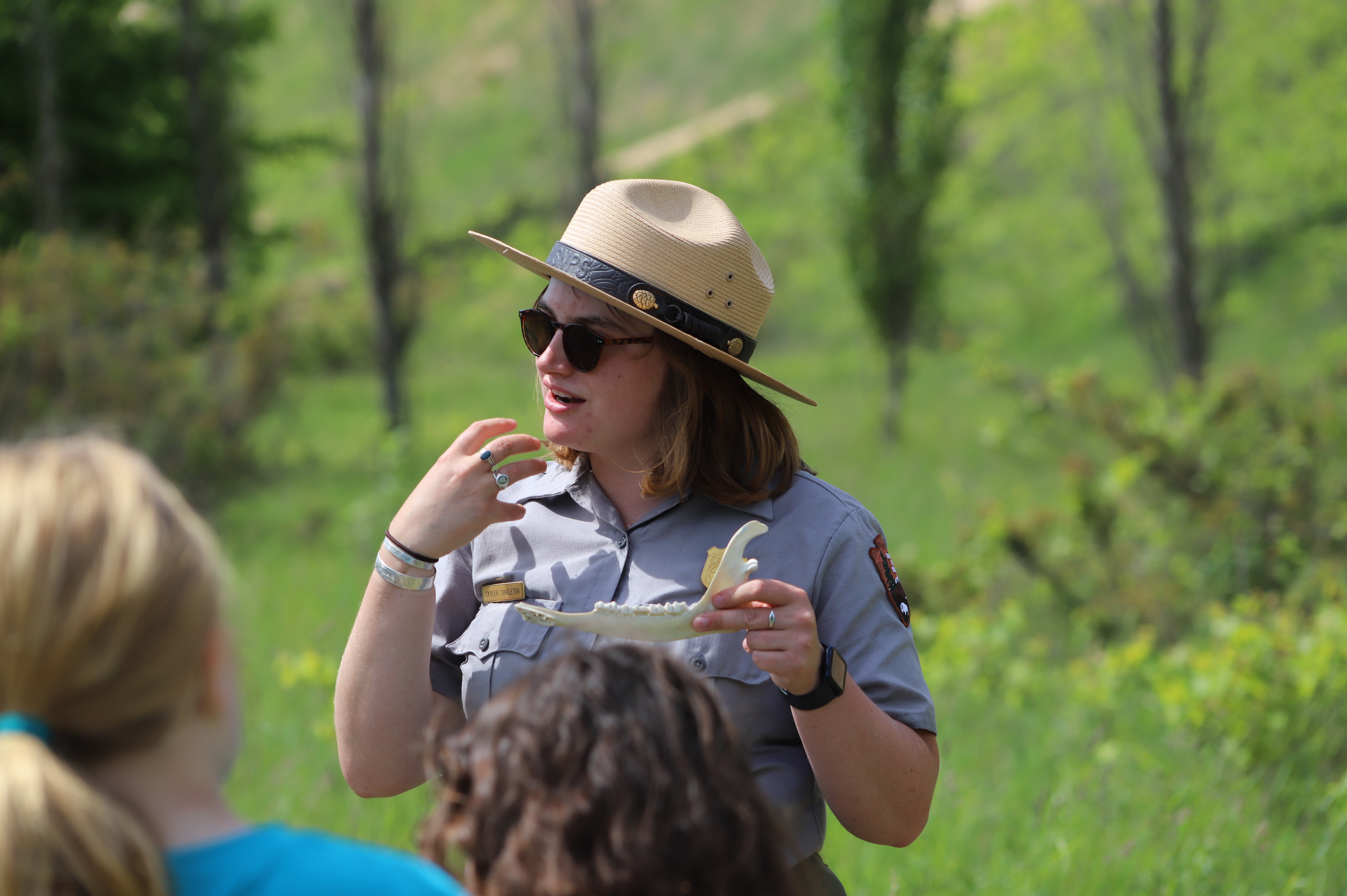 A ranger wearing a flat straw hat and sunglasses shows the jawbone of a deer to a group of students. The ranger is standing in a grassy field.