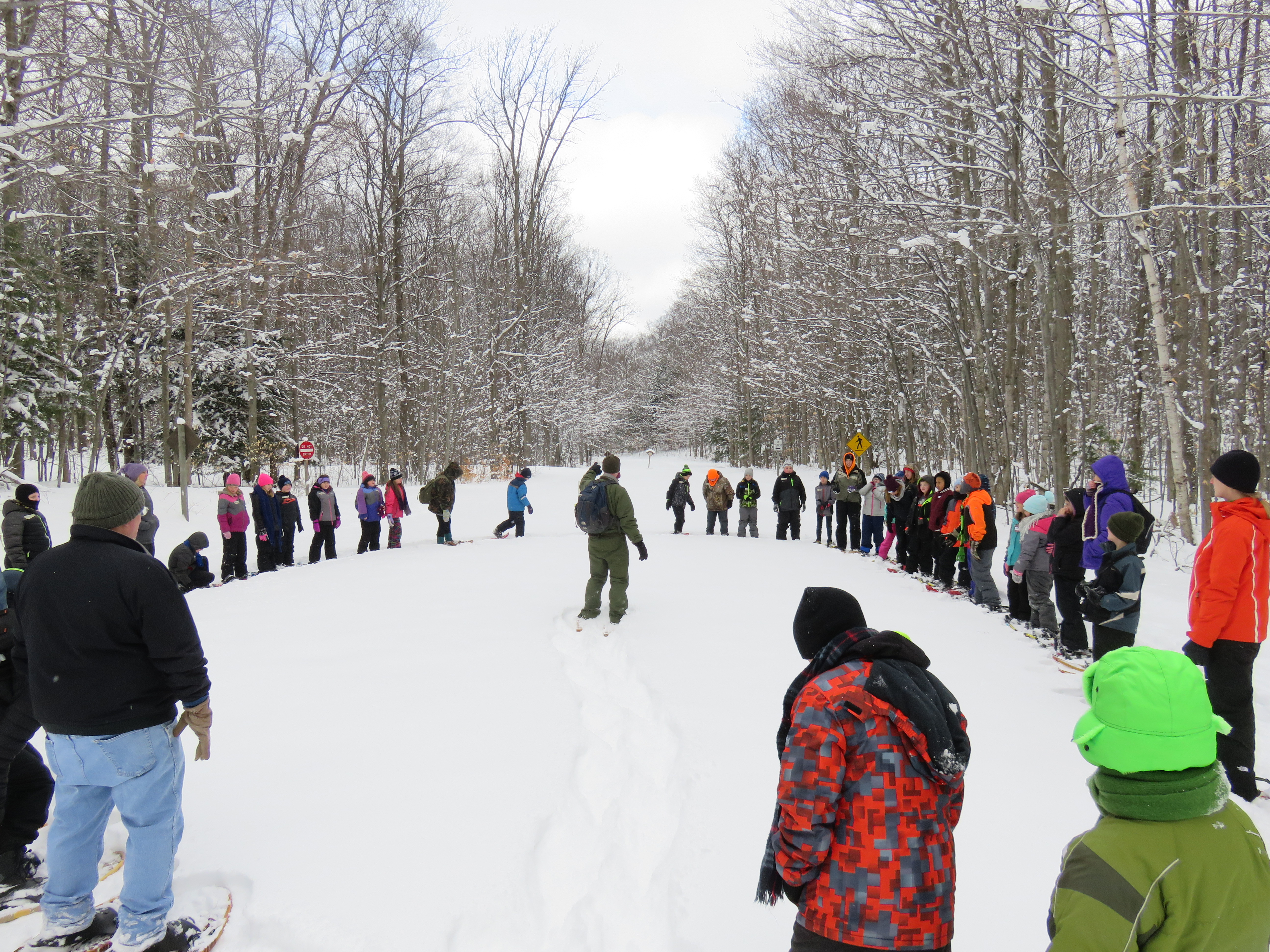 A group of students and chaperones wearing snowshoes stands in a circle in a snowy clearing around a ranger pointing towards a forested trail