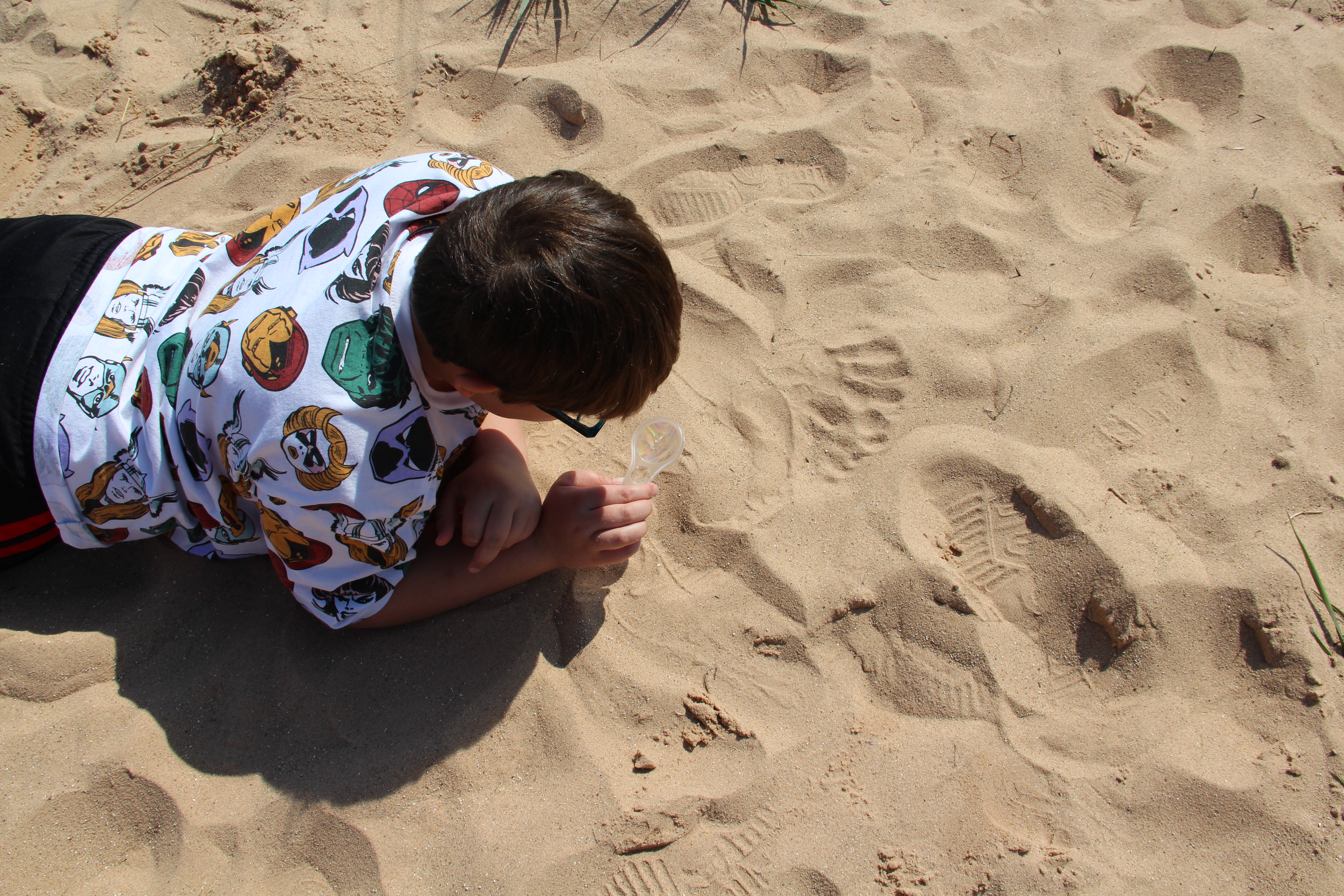 An elementary school student lays in the sand while using a magnifying glass to see closer