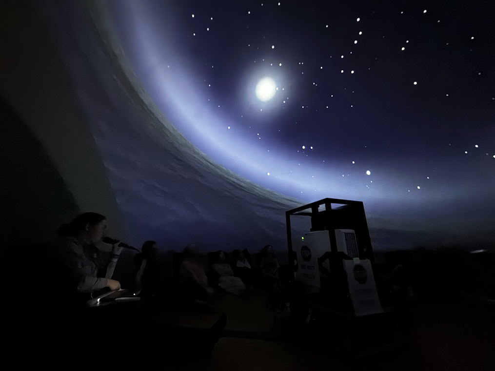 A ranger uses a microphone to speak to a group of students under a projected starry night sky