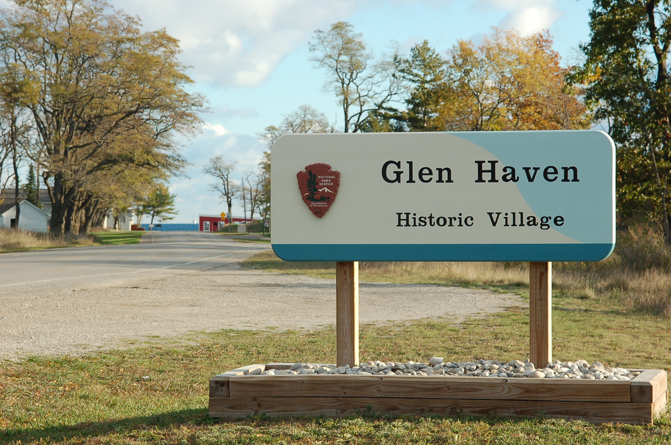 A sign with an NPS arrowhead symbol stands in front of a road lined with trees. The sign says "Glen Haven, Historic Village".