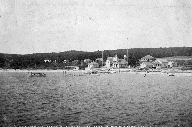 North Manitou Island Life Saving Station sits along the beach, including a boathouse and crew quarters, behind the station sits a row of cottages. All the buildings are painted white and stand out sharply from the dark forest in the background.