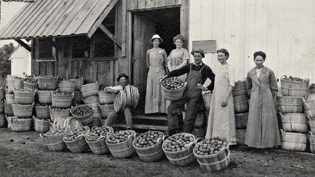 A family of farmers stands in front of a large wooden building, surrounded by baskets full of apples.